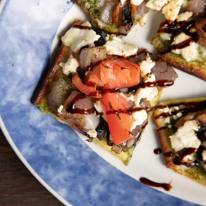 Close-up of flatbread with pesto, tomatoes, onions, feta, and balsamic glaze on a blue and white plate.