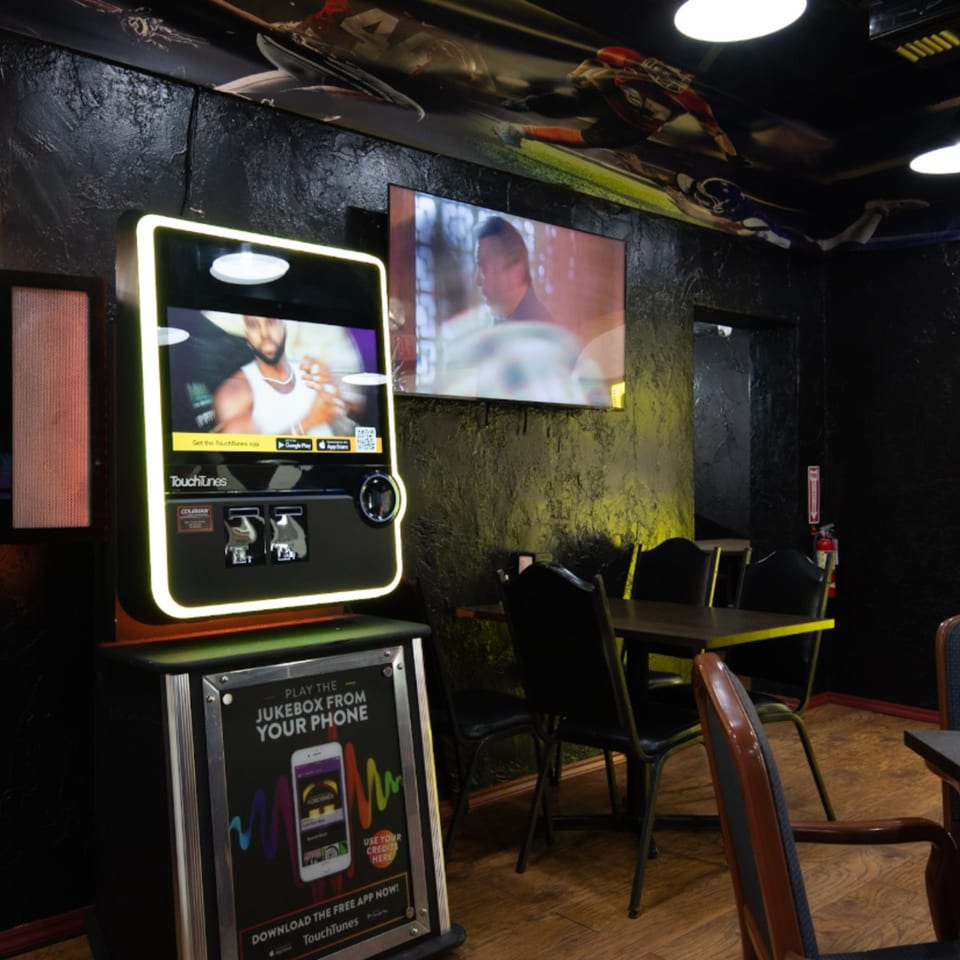 A jukebox and TV screen in a dimly lit sports bar with dark walls and tables.