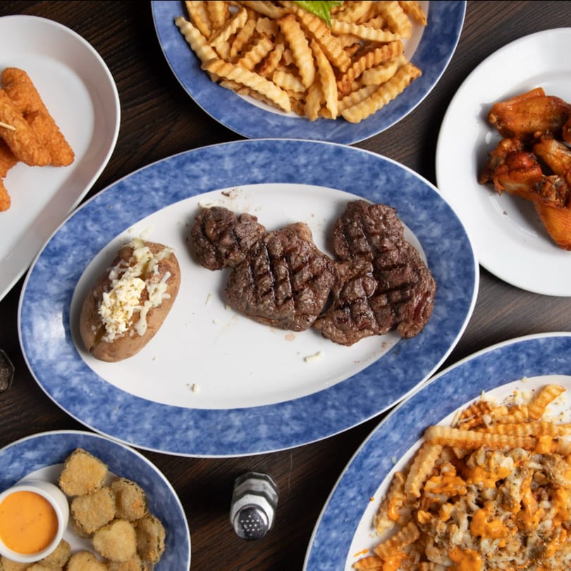 Overhead view of Game Time Sports Bar food: steak, baked potato, fries, wings, fried pickles, and chicken tenders.